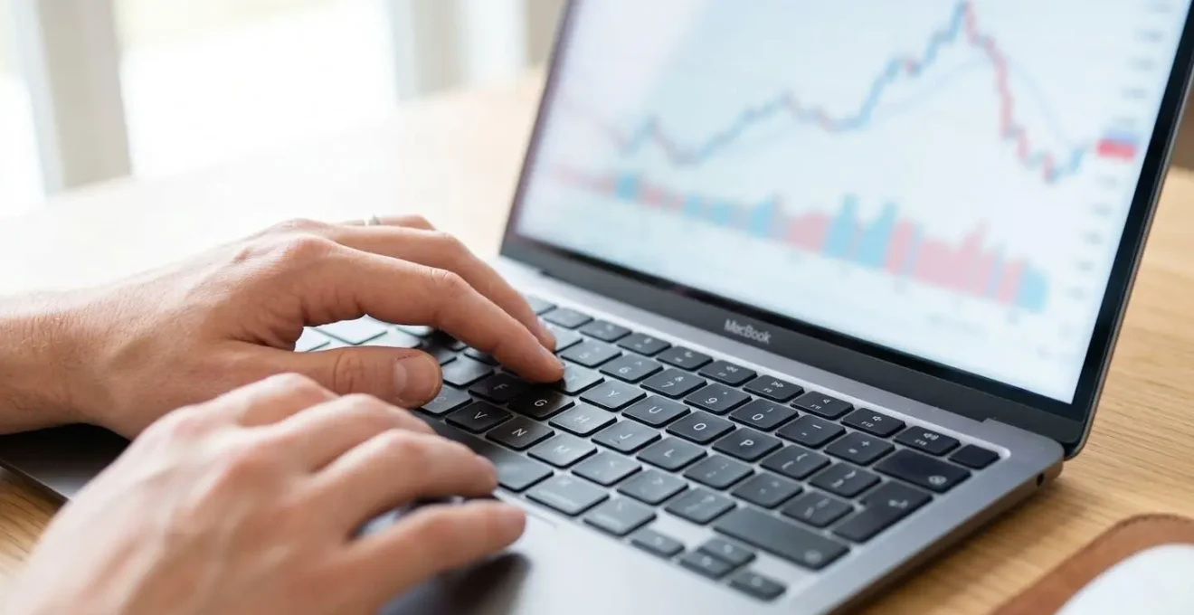 Gros plan sur des mains tapant sur un clavier d'ordinateur portable avec un graphique financier flouté visible à l'écran